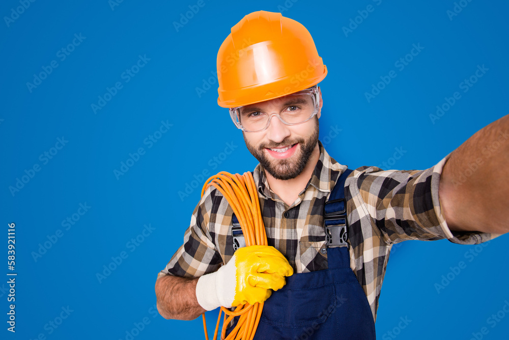 Self portrait of attractive joyful man in hardhat, overall, shirt, holding rolled cable on shoulder, blogger shooting selfie on front camera, isolated on grey background, having leisure Self portrait of attractive joyful man in hardhat, overall, shirt, holding rolled cable on shoulder, blogger shooting selfie on front camera, isolated on grey background, having leisure