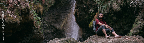 A young male tourist stands near a tropical waterfall among the rocks.