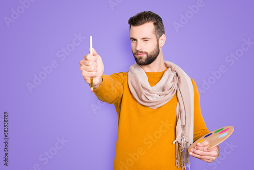 Portrait of creative concentrated artist with bristle in sweater and scarf on neck using, having colorful palette, brushed in arms, analyzing, expertising pic, isolated on grey background photo