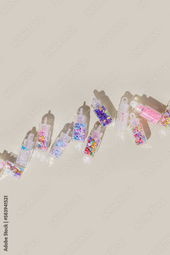 Colorful glitters diffrent form for nail art and makeup in small jars on beige background. Minimal aesthetic pattern with glitter for nails in glass bottles at sunlight, top view, flat lay