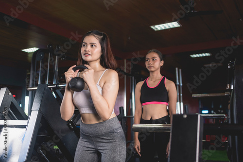 A young asian woman demonstrates how to do goblet squats to her friend. Training and teaching how to workout properly at the gym. © Mdv Edwards
