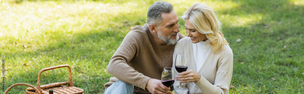 bearded middle aged man clinking glasses of wine with blonde and cheerful wife during picnic, banner. bearded middle aged man clinking glasses of wine with blonde and cheerful wife during picnic, banner.