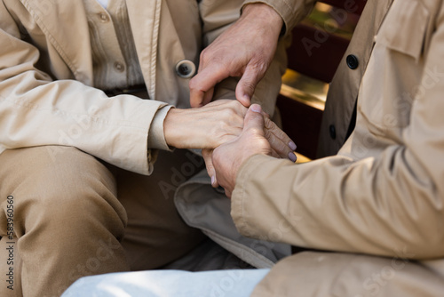 Cropped view of mature couple holding hands while sitting on bench in park.