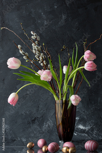 altrosa Tulpen mit Weidenkätzchen in stilvoller Vintage Glasvase. Umrahmt von goldenen und altrosa Ostereiern. Auf Glasplatte vor dunklem Hintergrund.
