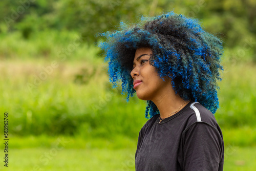 Foto de uma jovem negra de cabelos afro, tingidos de azul, com expressão serena no rosto, de perfil com a vegetação borrada do parque ao fundo