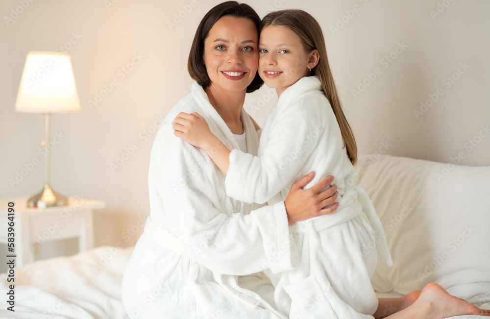 Beauty care at home together. Glad caucasian teen girl hugging young lady in bathrobe sit on white soft bed Beauty care at home together. Glad caucasian teen girl hugging young lady in bathrobe sit on white soft bed