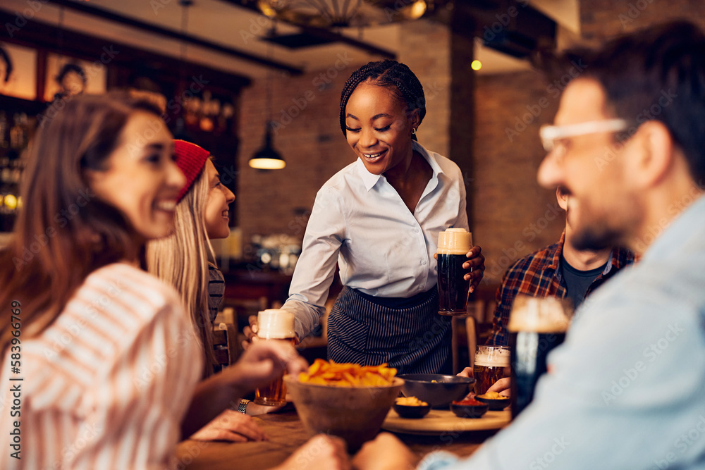 Happy black waitress serving beer to group of friends in pub.