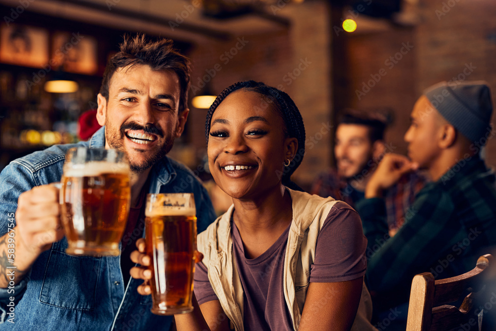 Happy multiracial couple toasting with beer in pub and looking at camera.