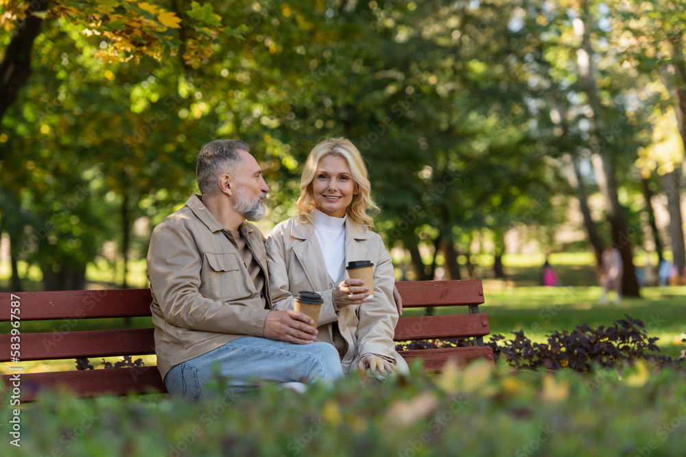 Carefree woman holding coffee to go near husband on bench in park. Carefree woman holding coffee to go near husband on bench in park.