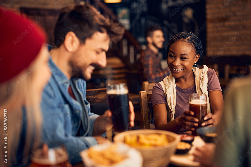 Happy black woman talks to her friends while drinking beer in pub.
