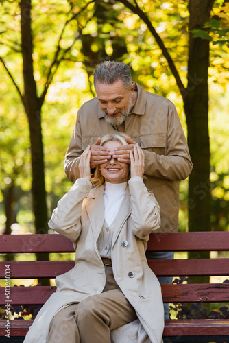 Positive mature man covering eyes of wife sitting on bench in park.