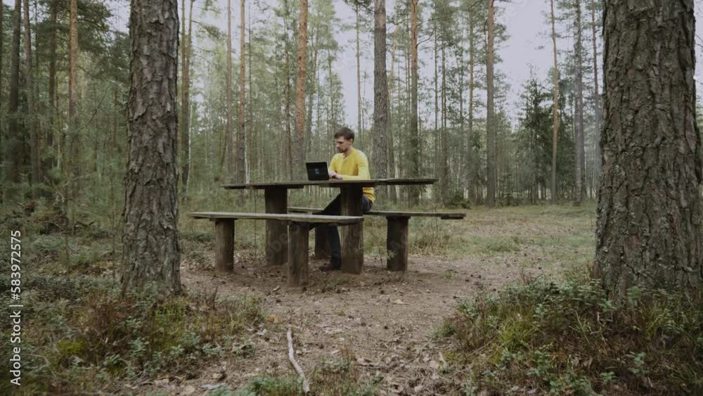 Thin man in yellow longsleeve works on black laptop in forest. Person is sitting on bench at wooden authentic table and typing on keyboard. Smooth stabilized camera approach to guy, wide shot. Thin man in yellow longsleeve works on black laptop in forest. Person is sitting on bench at wooden authentic table and typing on keyboard. Smooth stabilized camera approach to guy, wide shot.