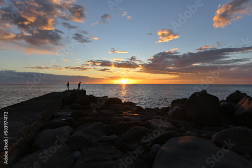 Ein schöner Sonnenuntergang über dem Meer und Angler stehen am Strand und Fischen © Fotolla