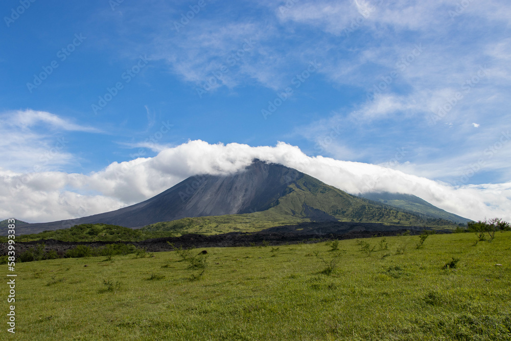 Pradera verde y lava seca con un volcán y un cerro con nubes en la cima. Cielo azul.