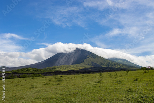 Pradera verde y lava seca con un volcán y un cerro con nubes en la cima. Cielo azul.
