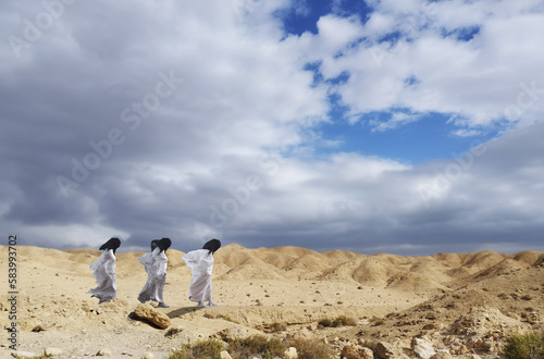 Three women in white capes and black headscarves walk against the backdrop of a sandy landscape with a blue sky.