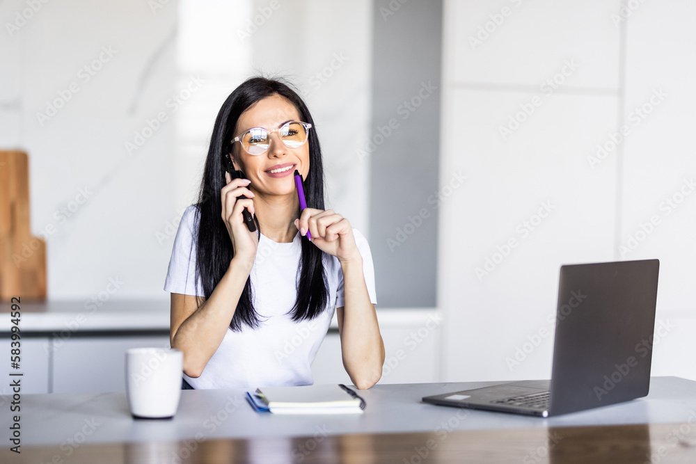 Portrait of a young woman talking the phone and working on laptop in kitchen
