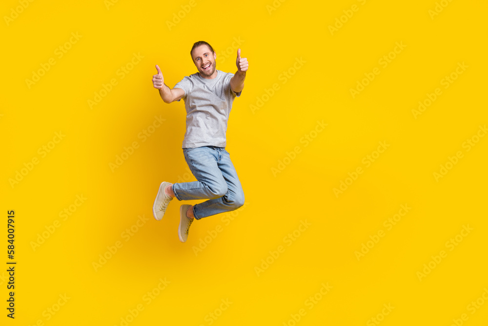 Full length photo of confident pretty man wear grey t-shirt jumping high thumbs up emtpy space isolated yellow color background