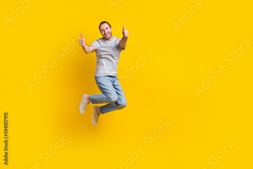 Full length photo of confident pretty man wear grey t-shirt jumping high thumbs up emtpy space isolated yellow color background
