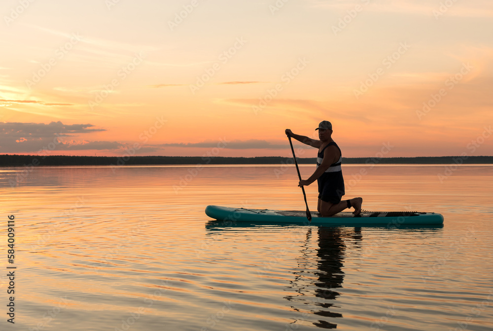 A man on his knees on a SUP board with an oar at sunset swims in the water of the lake against a pink sky reflection in the water.