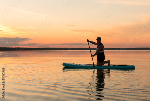 A man on his knees on a SUP board with an oar at sunset swims in the water of the lake against a pink sky reflection in the water.