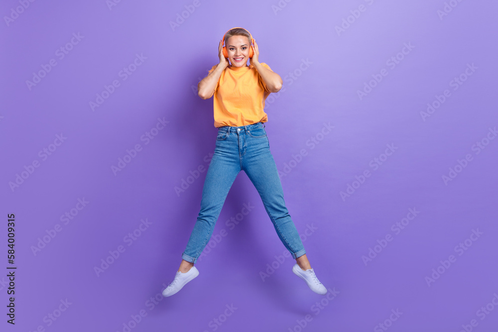 Full length photo of lovely cute girl dressed jeans white sneakers jumping hands touch headphones isolated on violet color background