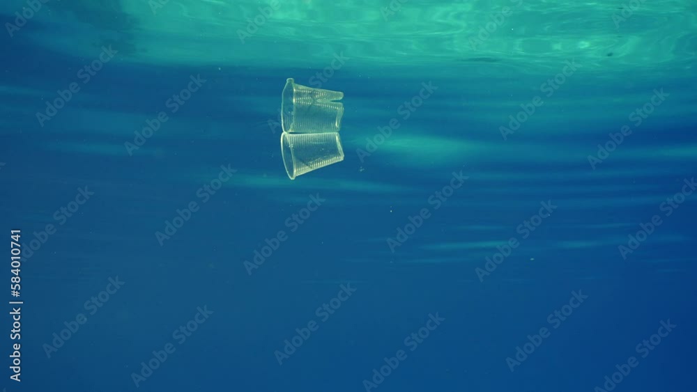 Plastic cup floating below surface o ocean, slow motion. Transparent disposable acrylic glass