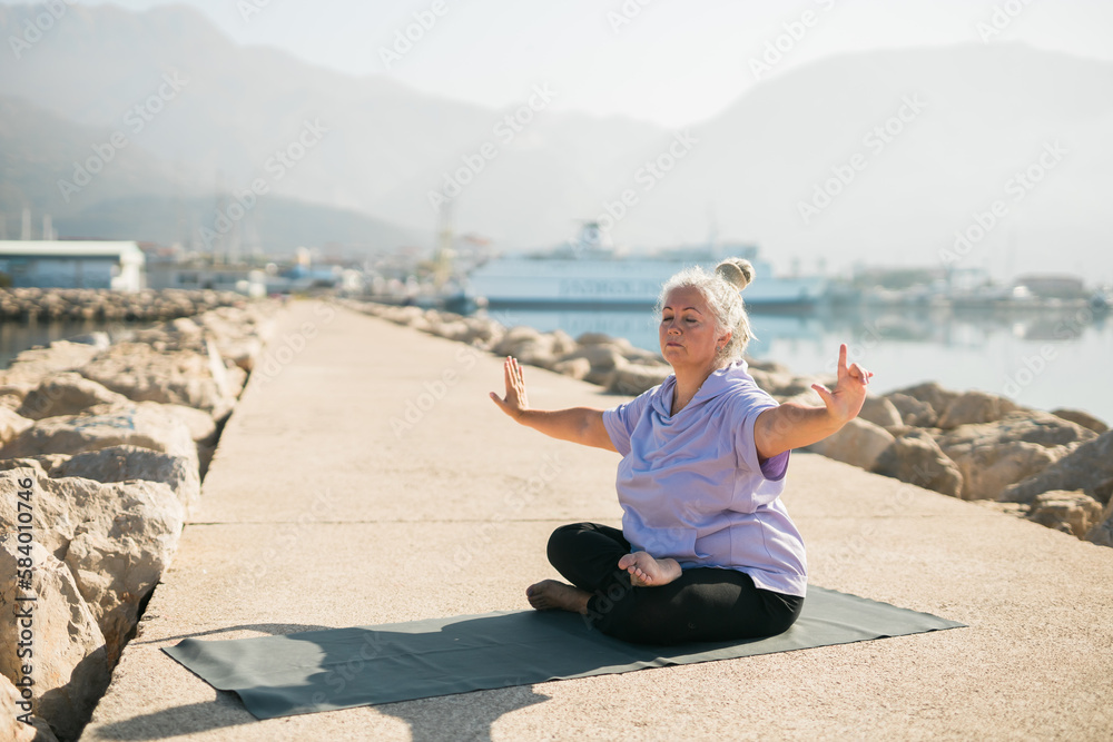Fototapeta premium Mature woman with dreadlocks working out doing yoga exercises on sea beach copy space - wellness well-being and active elderly age concept