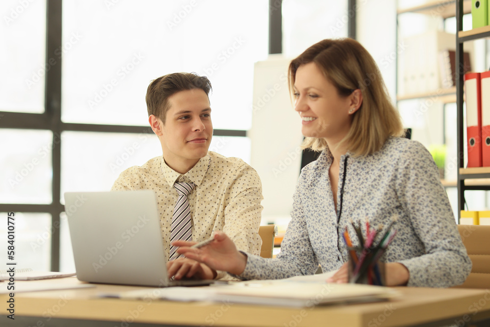 Colleagues sit at wooden table looking in laptop together Colleagues sit at wooden table looking in laptop together
