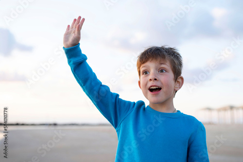 Surprised emotional child waving smiling, beach, sky with clouds in the background. Day light. Concept of travel, vacation, childhood, greeting. Caucasian boy 6 years old.