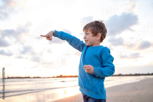 Surprised child stretched out his hand forward, pointing his finger, sea and the sky with clouds in background. Beach, sunset light. Concept of travel, vacation, childhood. Caucasian boy 6 years old.