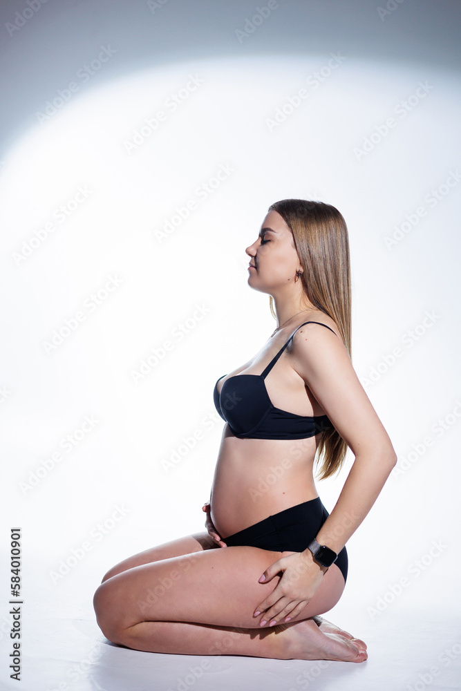 Fototapeta premium Cute pregnant woman with long hair in black underwear is sitting on a white background. Women's underwear for pregnant women.