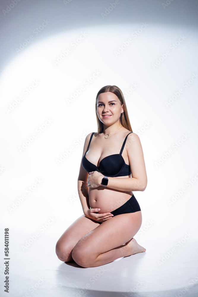 Obraz premium Cute pregnant woman with long hair in black underwear is sitting on a white background. Women's underwear for pregnant women.