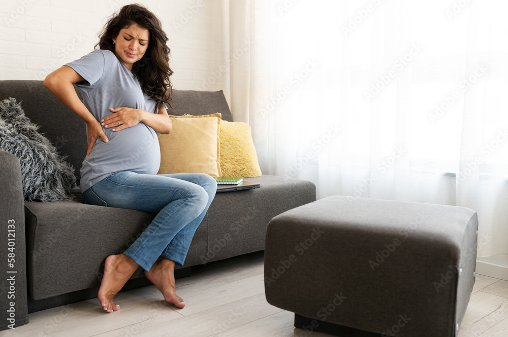 Beautiful 9 month pregnant woman sitting on the sofa at home having back pains caused by carrying a baby