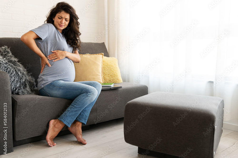Beautiful 9 month pregnant woman sitting on the sofa at home having back pains caused by carrying a baby Beautiful 9 month pregnant woman sitting on the sofa at home having back pains caused by carrying a baby