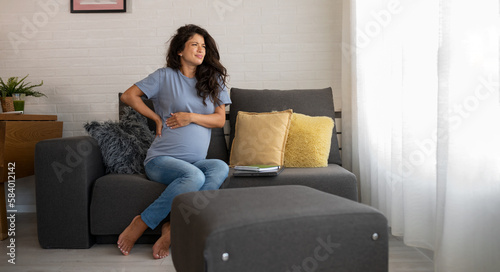 Free Download Beautiful 9 month pregnant woman sitting on the sofa at home having back pains caused by carrying a baby - GFXLoad