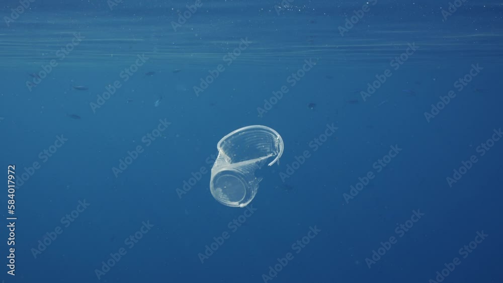 Disposable plastic cup floating under surface in blue water, slow motion. Transparent plastic