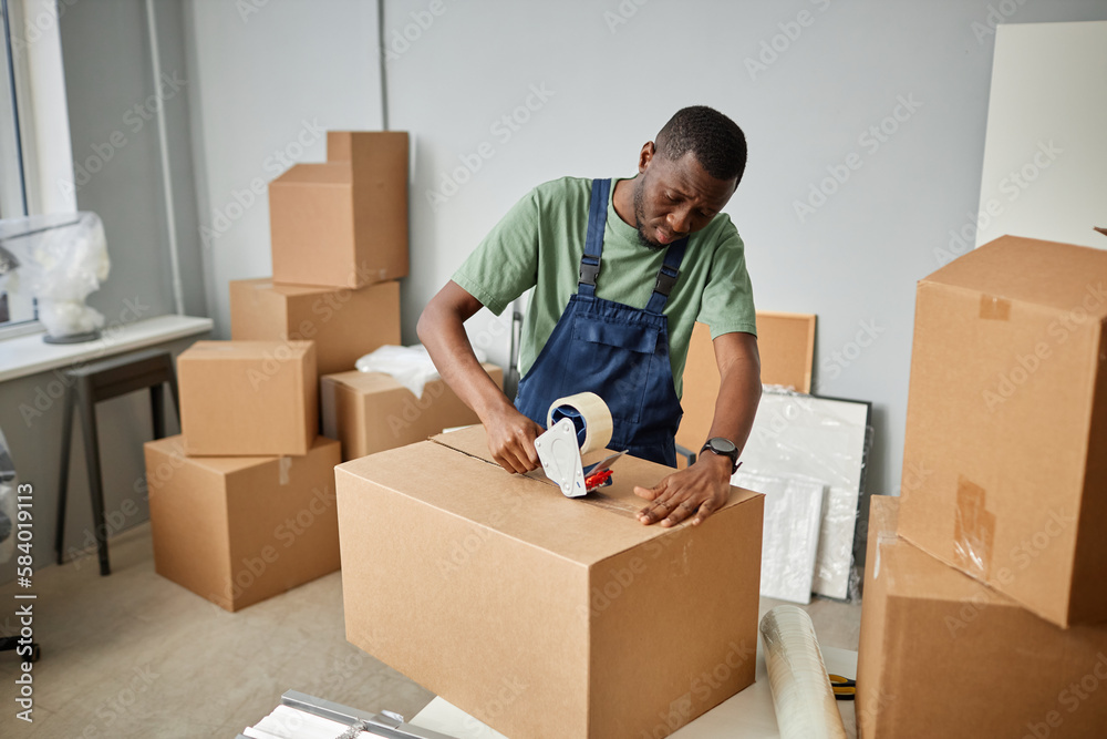 African American worker in uniform packing things in cardboard boxes with adhesive tape for relocation African American worker in uniform packing things in cardboard boxes with adhesive tape for relocation