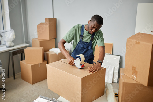 African American worker in uniform packing things in cardboard boxes with adhesive tape for relocation