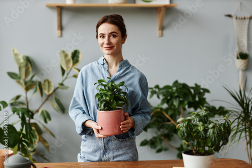 Waist up portrait of young woman posing with lush greenery at home and holding potted plant, looking at camera