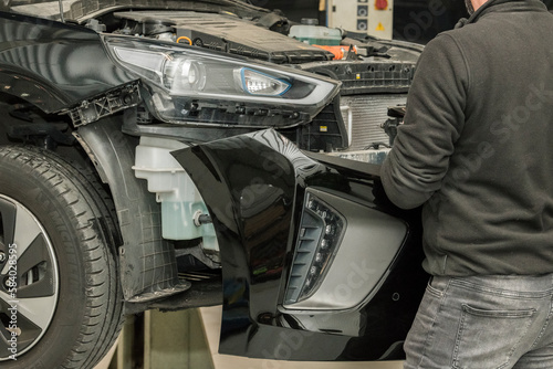 Detailed view of a blue-collar working mounting a bumper on the front of a black car. Automotive industry, maintenance, repair shop, garage workshop.