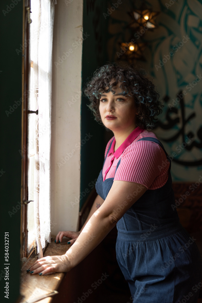 Portrait of a smiling brunette girl twenty-five years old dressed in a vintage pink blouse and blue velvet jumpsuit looks out the window in a cafe. Portrait of a smiling brunette girl twenty-five years old dressed in a vintage pink blouse and blue velvet jumpsuit looks out the window in a cafe.