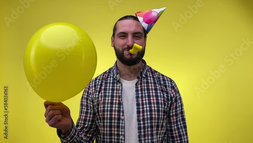 Cheerful mid adult man having fun wearing party hat blowing party horn, holding balloon, smiling isolated on yellow background. Bearded man congratulates sb. Happy birthday celebration holiday concept photo
