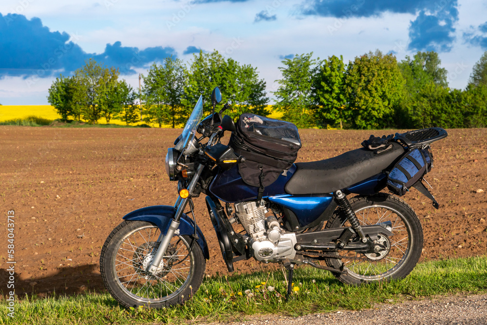 Fototapeta premium A motorcycle stands on the road against the background of a dug-up agricultural field on a sunny day. A motorcycle trip through nature. Motorcycle on a rainbow background.