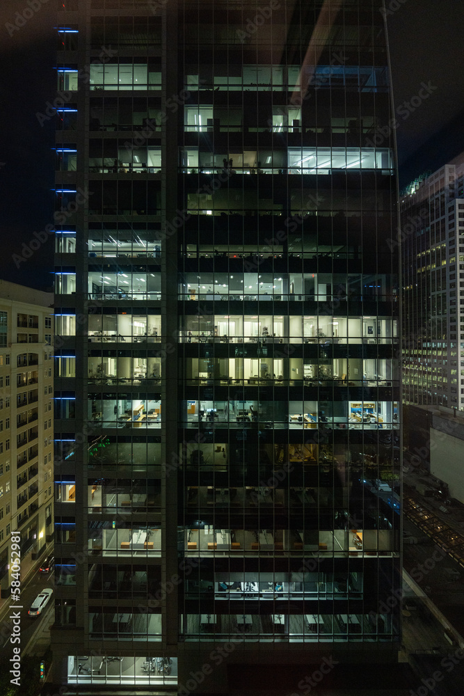 At night, the windows of a bustling office building light up to reveal the hardworking employees inside, dedicated to their tasks even after hours