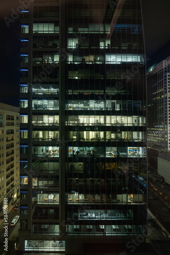 At night, the windows of a bustling office building light up to reveal the hardworking employees inside, dedicated to their tasks even after hours