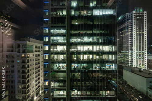 Night View of Busy Office Building: Illuminated Windows Reveal Dedicated Employees Working Late