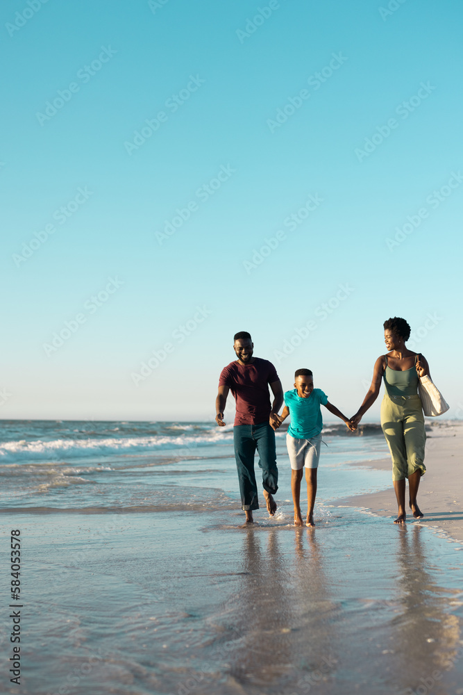Playful african american boy holding father and mother's hands and running in sea against sky
