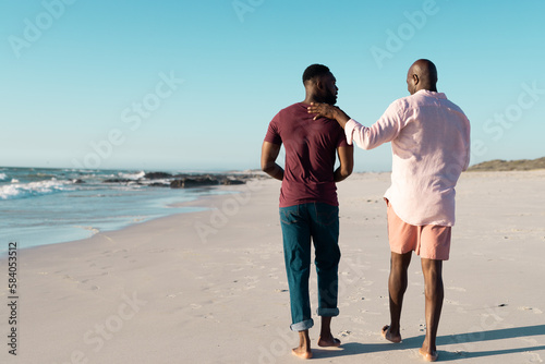 Rear view of african american mid adult son with senior father walking at beach against clear sky