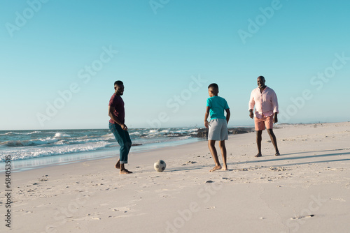 African american boy playing soccer with father and grandfather at beach against clear sky